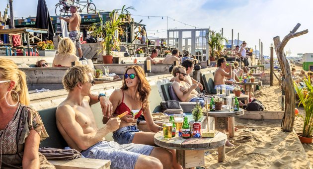 Een gezellig druk terras met mensen in badkleding met hun voeten in het zand bij strandtent Indigo op Scheveningen in Den Haag