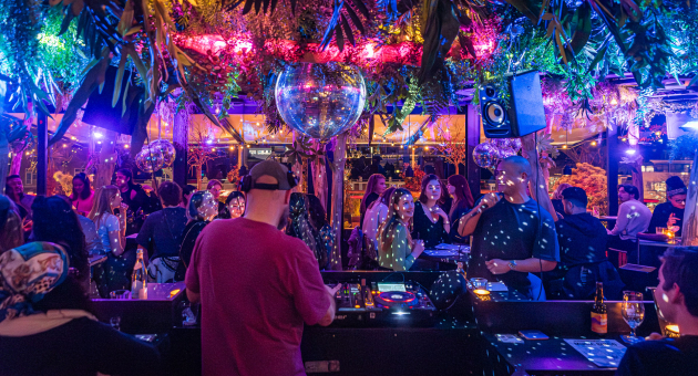 Een levendige nachtclub met een DJ, zingende man, discoverlichting, planten en dansende mensen
