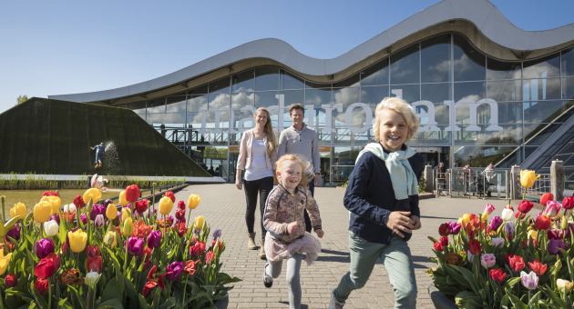 Een vrolijk gezin met twee kinderen loopt tussen kleurrijke tulpen richting de ingang van Madurodam, met een modern gebouw met golvend dak op de achtergrond onder een heldere blauwe lucht.