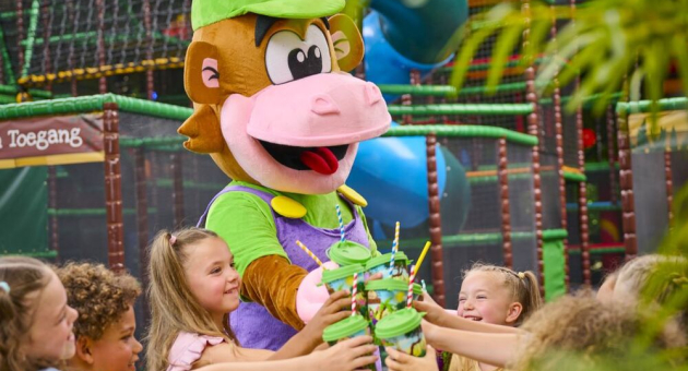 Een vrolijke mascotte in de vorm van een aap deelt bekers uit aan lachende kinderen in een kleurrijke indoor speeltuin.