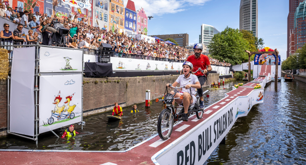 Twee deelnemers op een tandemfiets racen over een schans tijdens Red Bull Showrun in een drukbezocht, kleurrijk Den Haag.