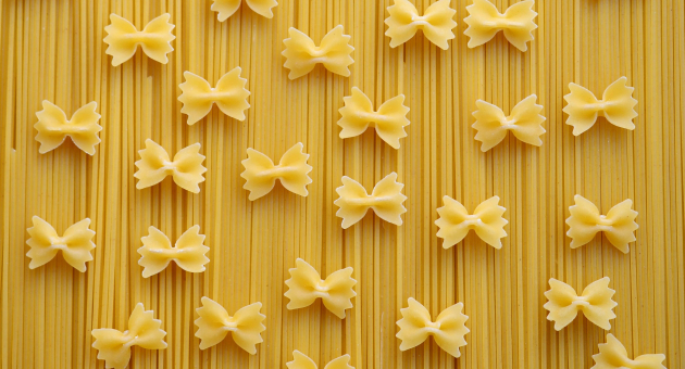 Droge strikvormige pasta (farfalle) ligt verspreid over een ondergrond van rechte ongekookte spaghetti, in een patroon tegen een gele achtergrond.