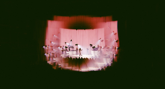 Eefje de Visser op het podium in een dromerige, roze-gloeiende setting, omringd door muzikanten en sfeervolle verlichting. Het beeld straalt een mysterieuze en filmische sfeer uit, perfect passend bij haar kenmerkende livebeleving.