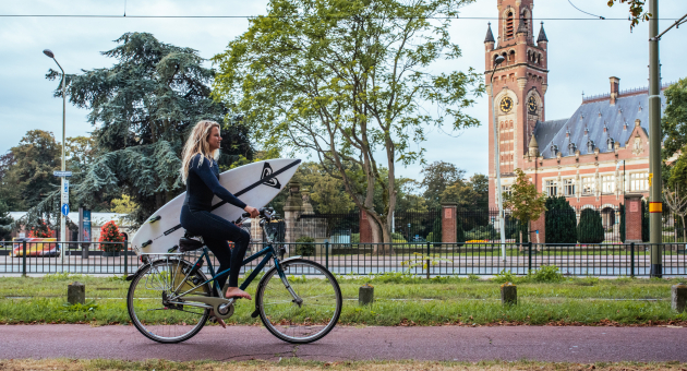 Zomer in Den Haag. Meisje met surfplank onder haar arm fiets langs het Vredespaleis richting Scheveningen