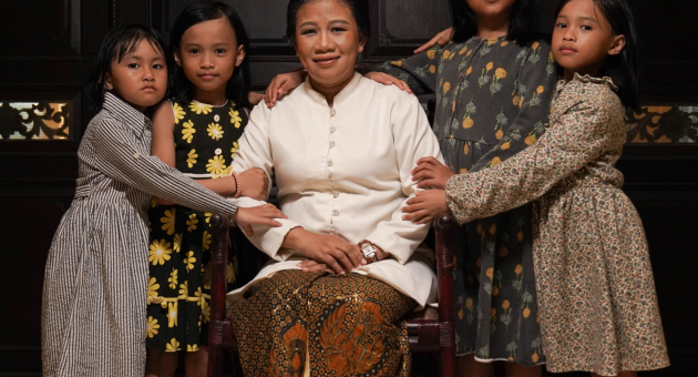 Een vrouw in traditionele kleding zit omringd door vijf jonge meisjes in jurken, die haar liefdevol vasthouden.