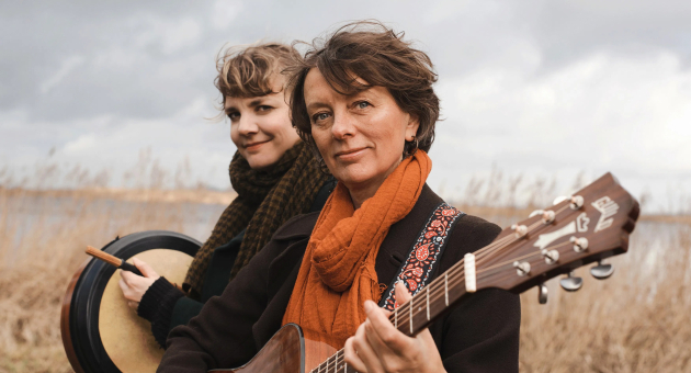 Twee muzikanten buiten bij riet en water, één met gitaar en oranje sjaal, de ander met een trommel en stok.
