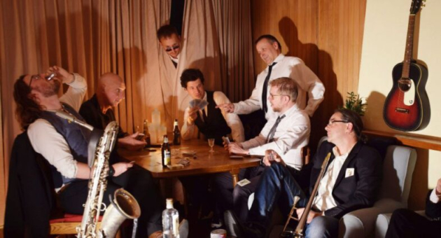 De band in een setting vol rock-’n-roll en jaren 20-swing met kaartspel, drankje en gitaar aan de muur.