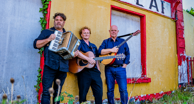 Drie mannen met accordeon, gitaar en bouzouki poseren voor een vervallen geel-rood gebouw met het woord "CAF" op de gevel.