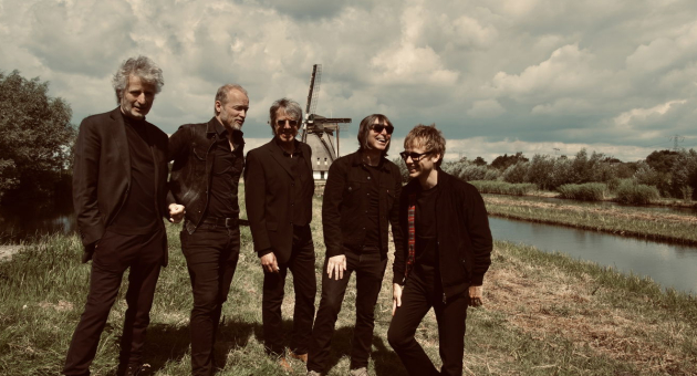 De leden van coverband Timebox poseren lachend in zwarte outfits op een dijk met een molen op de achtergrond.