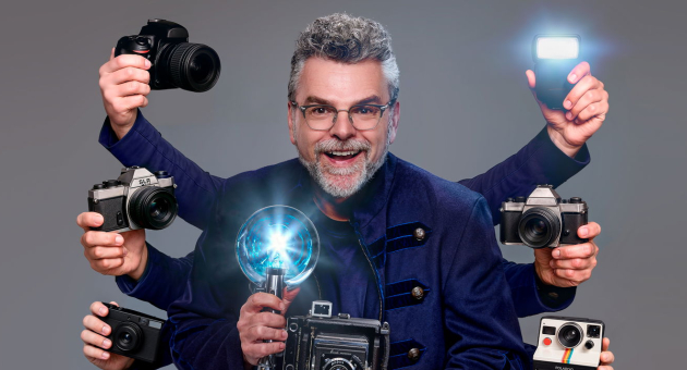 William Rutten staat lachend voor een grijze achtergrond. Hij heeft 7 armen met verschillende cameratoestellen in zijn handen en draagt een donkerblauwe jas.  