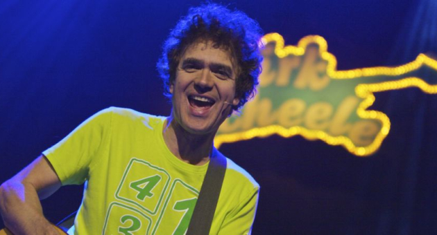 Dirk Scheele vrolijk en spelend op gitaar op podium in geel shirt en geel bandlogo met zijn naam erin passend bij zijn kindervoorstelling popconcert Foto: Guido Breedveld