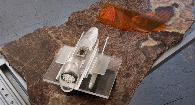 Kunstwerk op een natuurstenen blad met een transparant, mechanisch ogend object en een oranje plexiglazen vorm, geplaatst op een metalen onderstel. 