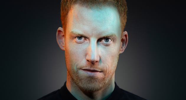 Close-up van René van Meurs met een serieuze blik. Hij draagt een zwart shirt en staat voor een zwarte achtergrond. Zijn gezicht wordt belicht met oranje en blauw licht.