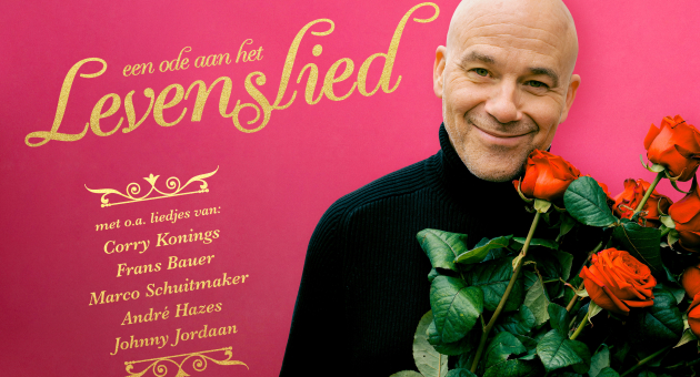 Chris Grem draagt zwarte coltrui. Hij heeft een bos rozen vast en kijkt lachend in de camera. 