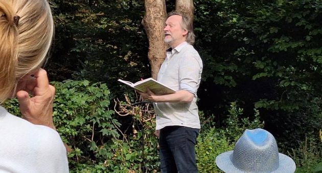Man leest voor uit een boek tijdens een buitenoptreden in een groene, bosrijke omgeving; toeschouwers luisteren vanaf stoelen op het gras.