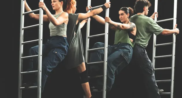 Vier dansers bewegen tussen metalen klimrekken in een choreografie op een donker podium