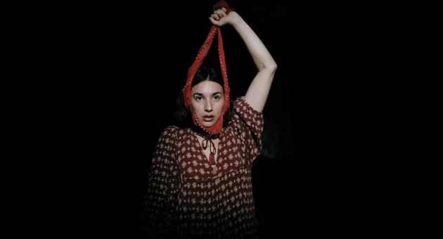 Promotiebeeld dansclick Melisa Diktaş in een rode bloemenjurk kijkt recht in de camera, met een rode stoffen band strak rond haar gezicht en omhooggehouden arm, tegen een donkere achtergrond.
