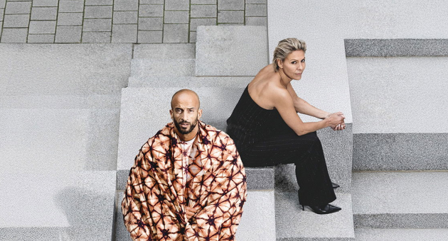 Foto van Mouna en Walid van schuin bovenaf. Ze zitten op grijze betonnen blokken en kijken recht in de camera. Mouna draagt een zwarte jumpsuit en khalid een rood-oranje tie-dye jas. 