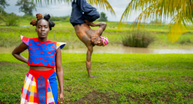 Afbeelding in groen achtige omgeving met een danser die op 1 hand staat en een meisje op de voorgrond in kleurrijke kleding met haar hand in der zij. 