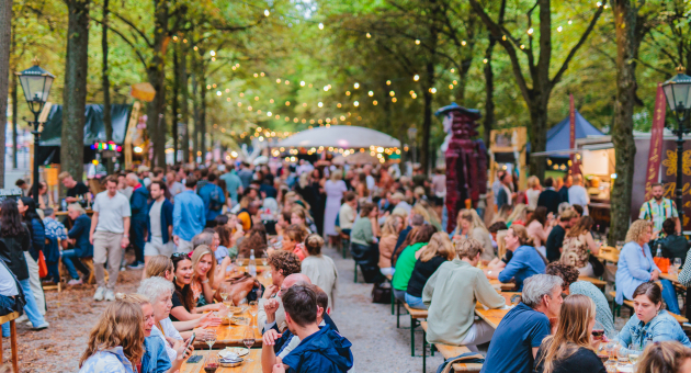 Drukke markt met lange picknicktafels onder bomen, mensen eten en drinken onder sfeerverlichting