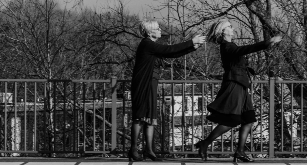 Zwart-wit promotiebeeld voorstelling dubbelspoor: Twee oudere vrouwen met witgrijs haar bevinden zich op het perron van een treinstation. Een van de vrouwen glipt weg terwijl de andere vrouw haar armen naar haar toereikt om haar te vangen.