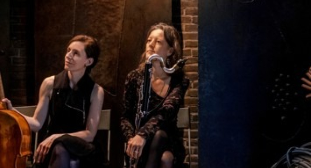 Katharina Gross en Fie Schouten zitten naast elkaar op het podium met hun instrumenten: de cello en de basklarinet. Ze dragen donkere kleding en kijken geamuseerd naar de linkerkant. 