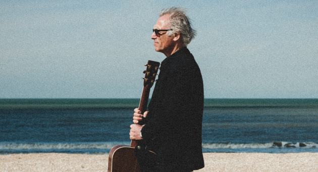 Philip Kroonenberg met gitaar in de hand op het strand, in de wind kijkend met zonnebril wat past bij emotie van zijn country genre.