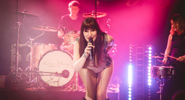 De leden van de Taylor Swift Tribute Band staan op een roze verlicht podium. De zangeres kijkt lachend de camera in. 