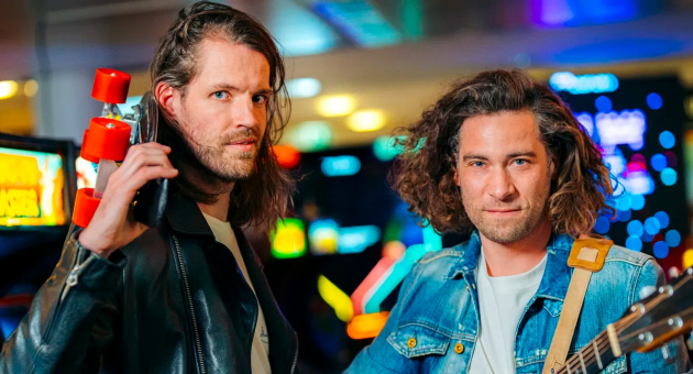 De leden van Popup choir, Maarten Bos en gitarist Tijmen de Koning, staan met serieuze blikken in een ruimte vol arcade machine's.  