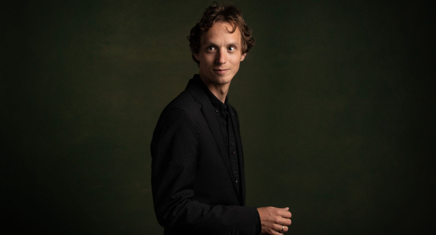 Jonge man met krullend haar in een zwarte outfit kijkt opzij en poseert tegen een donkere, groen getinte achtergrond.