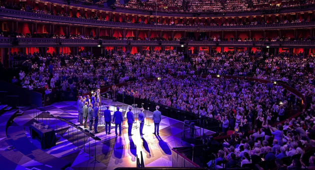 Een groep mensen staat op een groot podium in een volle concertzaal met sfeervolle paarse verlichting.