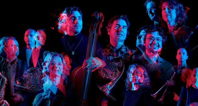 Groep muzikanten in kleurrijk blauw-rood licht, sommigen met blaasinstrumenten of een cello