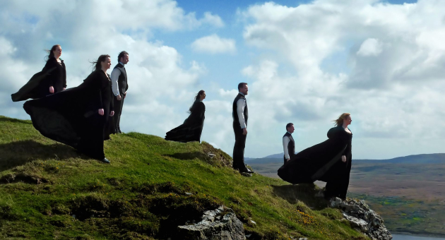 Zanggroep in zwarte gewaden op een groene klif, met wapperende capes in de wind en een weids, heuvelachtig landschap op de achtergrond.