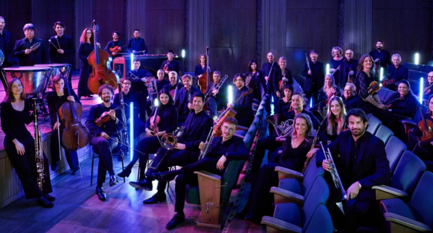 Leden van het WDR Funkhausorchester zitten met hun instrumenten op stoelen in een theaterzaal