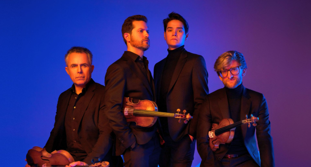 Vier mannen in zwarte pakken met strijkinstrumenten, poserend tegen een blauw-paars verlichte achtergrond