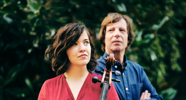 Twee mensen staan buiten voor een groene achtergrond; de vrouw vooraan houdt een cello vast, de man staat erachter.