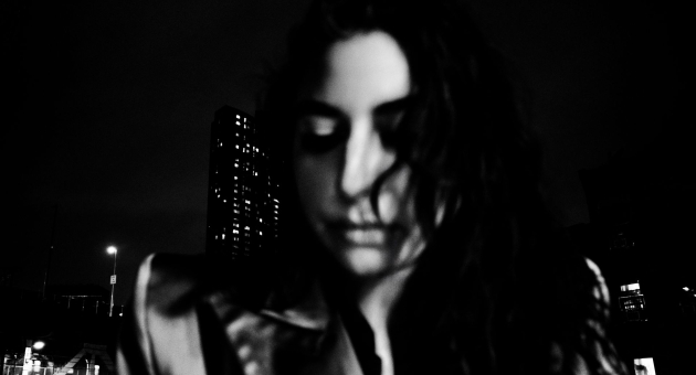 Zwart-wit portret van een vrouw met loshangend haar die naar beneden kijkt, met op de achtergrond een nachtelijk stadslandschap en een flatgebouw met brandende lichten.