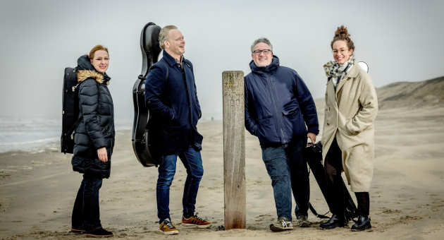  Vier musici poseren op een winderig strand, gekleed in warme jassen. Twee van hen dragen een cellokoffer op de rug. Ze staan ontspannen rond een houten paal in het zand, met duinen op de achtergrond en de zee in de verte.