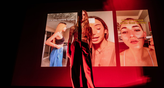 Een vrouw staat op een podium in rood licht, met projecties van drie verticale video’s van influencers op de achtergrond die schoonheidsproducten laten zien. Haar silhouet mengt met de beelden.