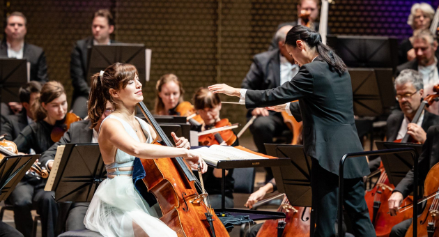 Dirigent leidt een orkest terwijl een celliste een solo speelt tijdens een concert.