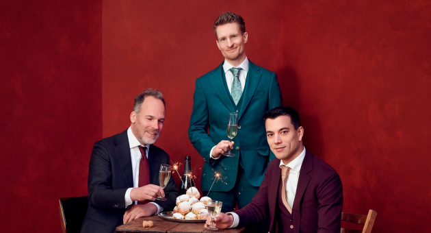 Joep Hullegie en Andries Tunru zitten aan een tafel met oliebollen, champagne en sterretjes. Stefan Hendrikx staat aan het hoofd van de tafel. De mannen zijn gekleed in pakken met stropdassen en staan voor een rode achtergrond. 