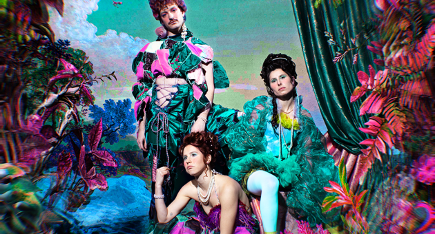 Drie performers van Collectief Aqueerius in extravagante barok-wave kostuums poseren in een kleurrijke, fantasierijke setting. De visuele stijl is een mix van barok en popart
