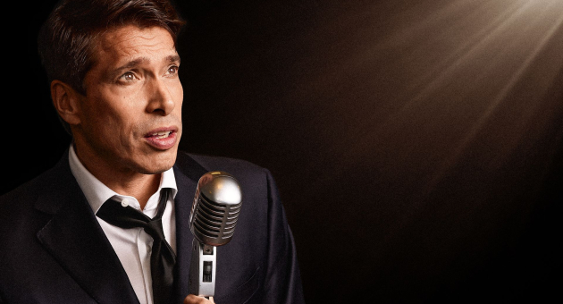 Man in pak met stropdas spreekt in een oude microfoon, spotlicht op hem gericht.