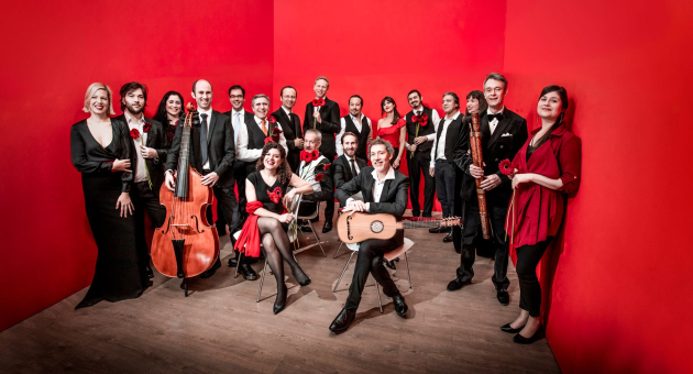 Een groot muziekensemble poseert in een felrode kamer met houten vloer. De groep bestaat uit mannen en vrouwen in feestelijke zwart-rode kleding, met diverse instrumenten zoals cello, gitaar, fluit en viool. Ze staan en zitten in een losse formatie en lachen naar de camera.
