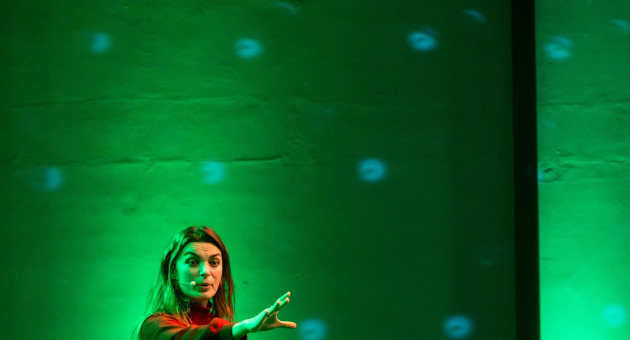 Vrouw op een groen verlichte podiumvloer tijdens een presentatie of theatervoorstelling.