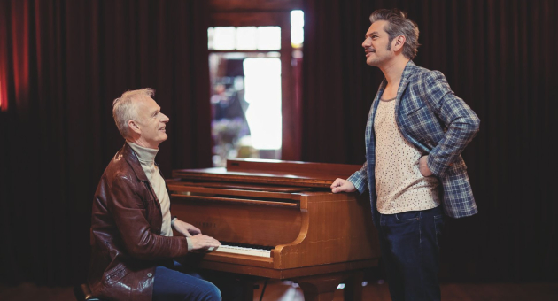 Twee mannen bij een houten piano, één spelend en de ander staand en lachend in gesprek