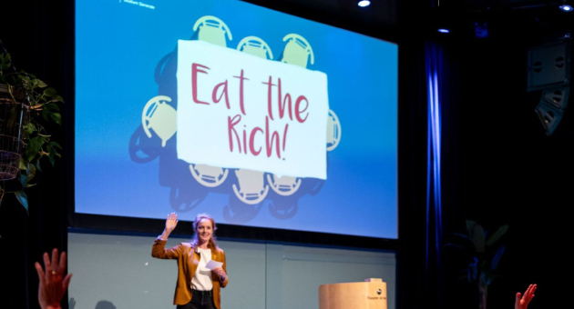Spreker op podium voor groot scherm met tekst “Eat the Rich”, publiek steekt handen op in zaalsetting