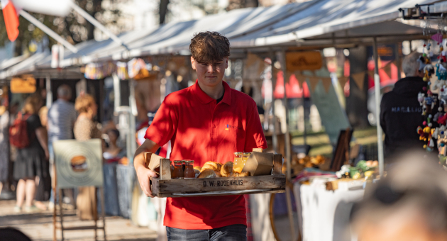 Een jongen in een rode polo draagt een dienblad met eten op een markt.