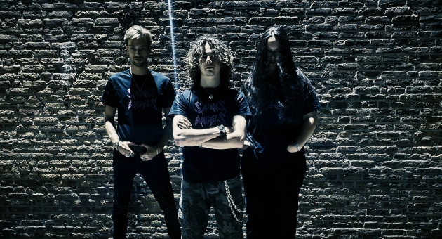 Drie mannen in donkere kleding poseren ’s avonds voor een bakstenen muur