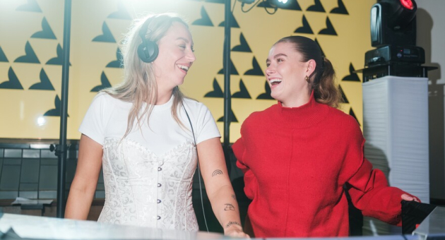 Twee lachende meisjes achter een dj tafel. Eén heeft een koptelefoon op. 